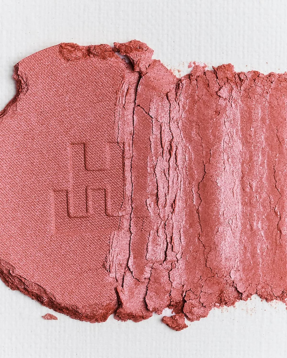 Crushed beige-pink pressed powder with FH embossing smeared across textured white paper, showing a satin sheen