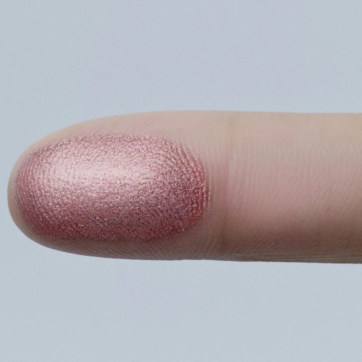 Fingertip swatch of a sparkly metallic rose pink eyeshadow showing fine shimmer against a pale blue background.