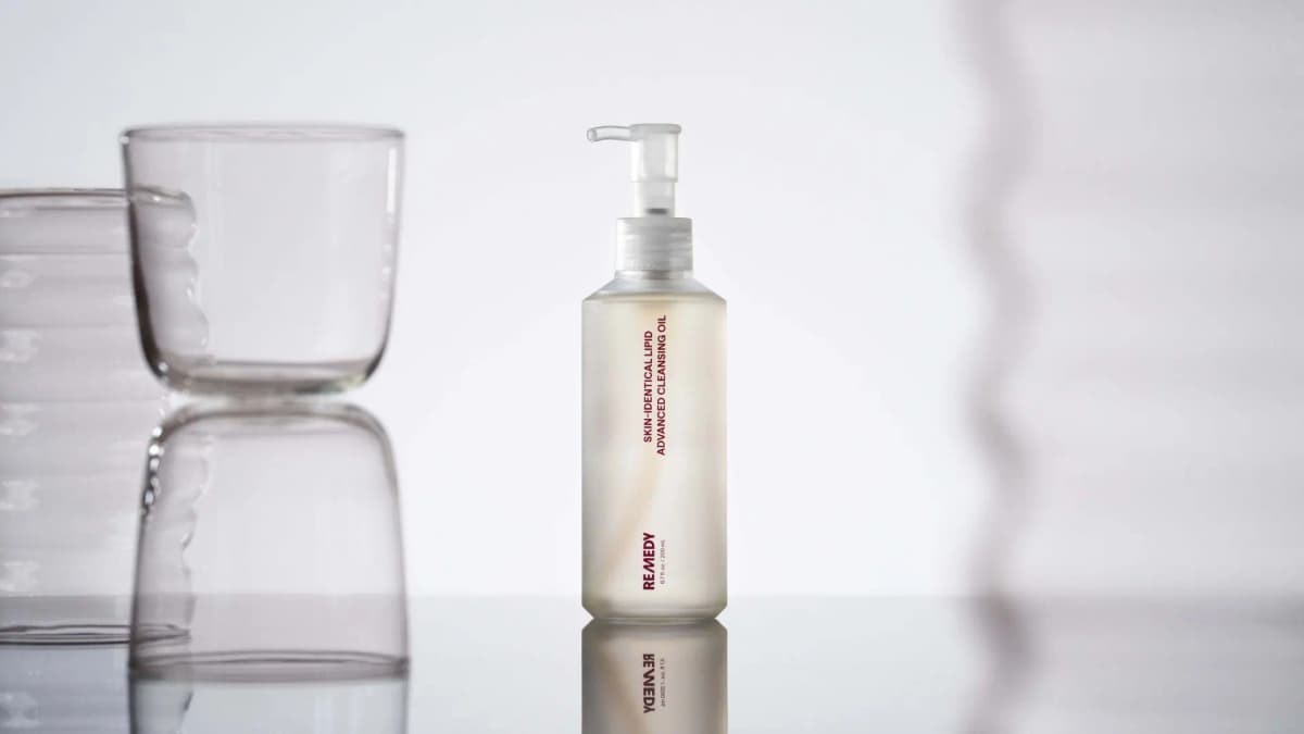 Remedy Science Skin-Identical Lipid Advanced Cleansing Oil frosted pump bottle on a reflective surface beside clear glassware