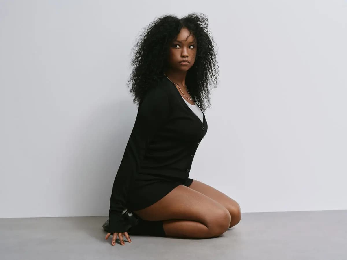 Model with voluminous black curly hair kneeling on a grey floor in a black ribbed cardigan, glancing upward