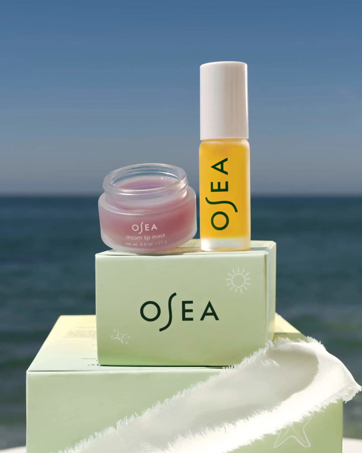 Open pink dream lip mask jar and yellow lip oil tube on mint green OSEA boxes against the sea