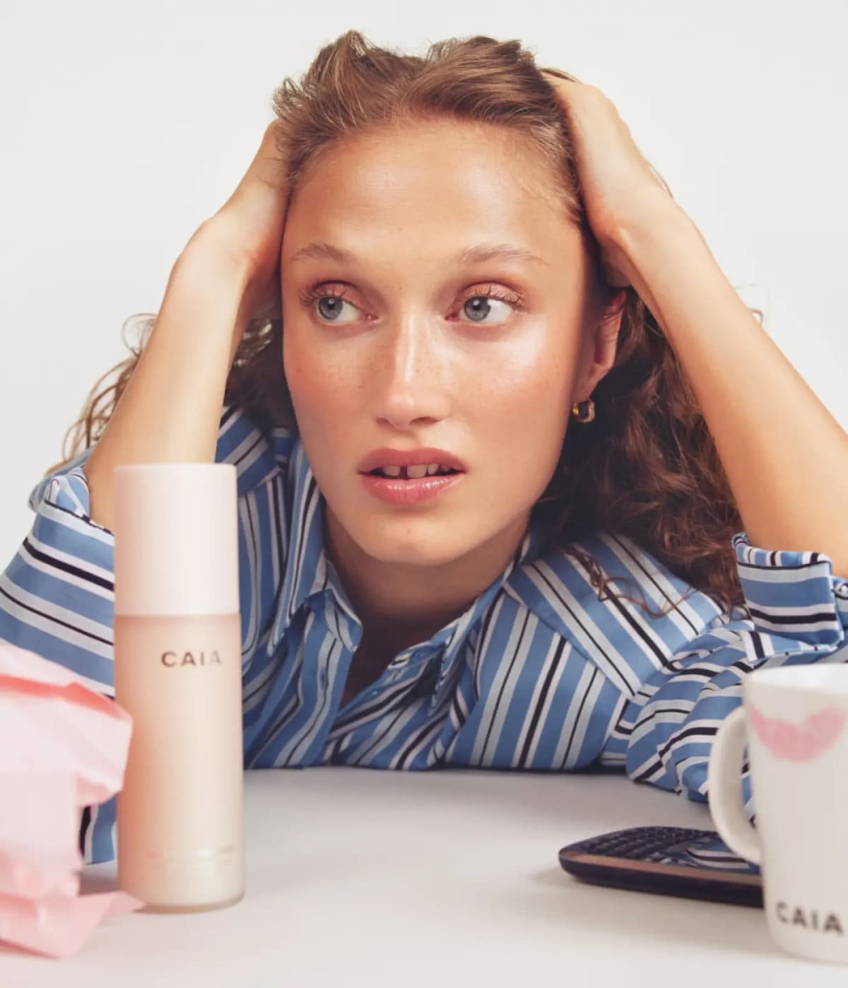 Freckled woman in a blue striped shirt rests her head in her hands behind a CAIA spray bottle and CAIA mug