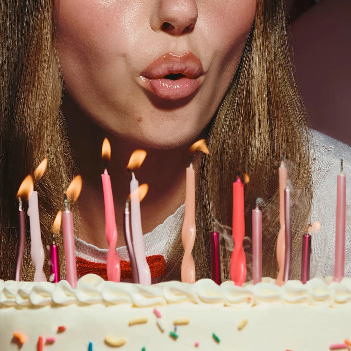 Close-up of glossy, shimmery nude lips blowing out pink candles on a white frosted sprinkle cake