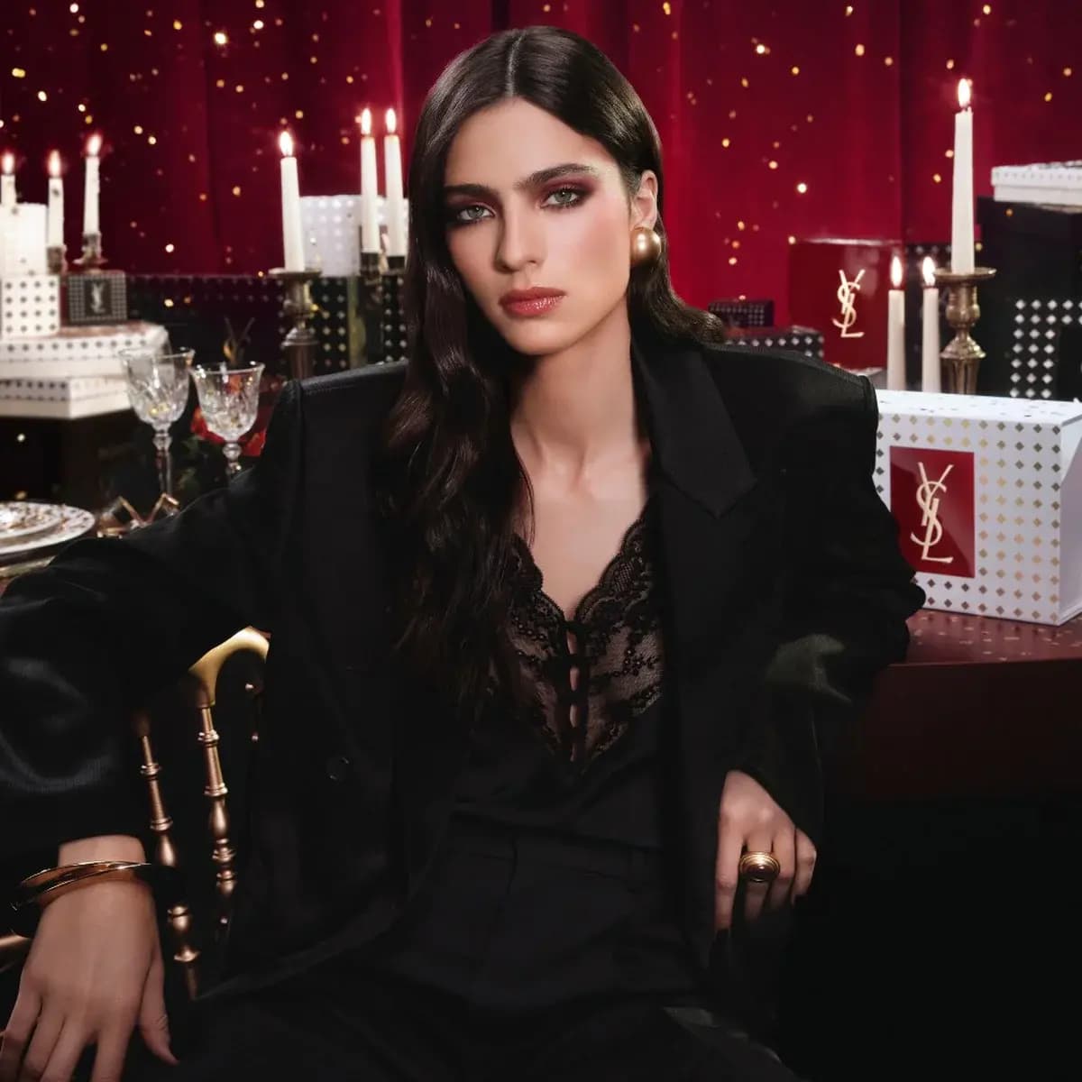 Model in black suit and lace top with smoky burgundy eyes, seated before candles, red curtain and YSL gift boxes
