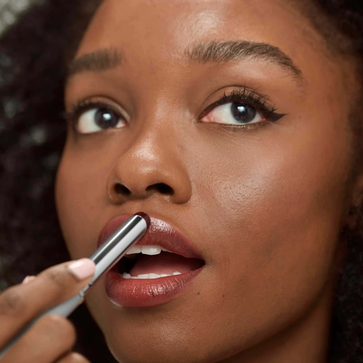 Woman with deep skin tone applying the dark berry lipstick from a silver tube, lips glossy plum-brown