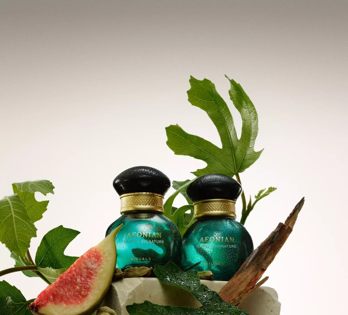 Two dewy teal Rituals Aeonian Santal Signature bottles with fig leaves, a fig wedge, cardamom pods and sandalwood