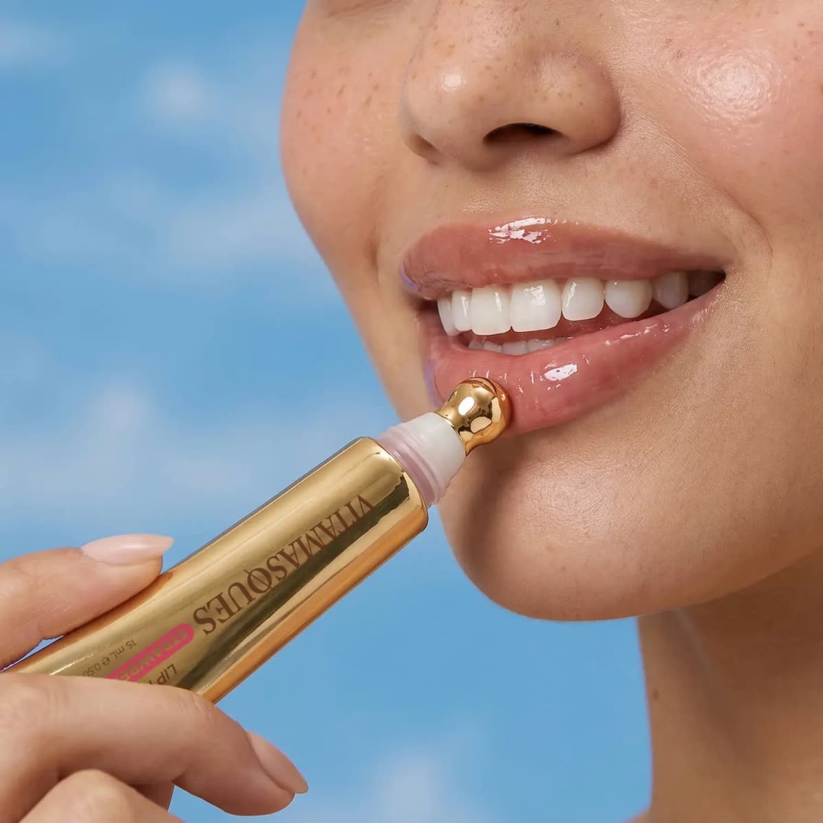 Smiling model applying the glossy pink tint with the tube's gold ball-tip applicator, blue sky behind