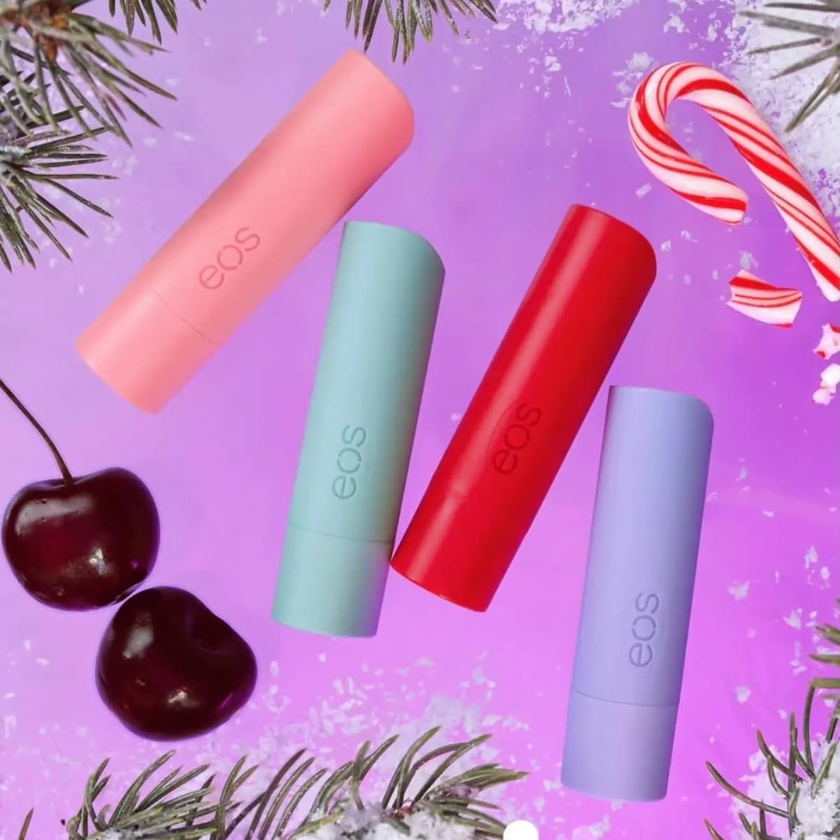 Pink, mint, red and lavender eos lip balm sticks on a purple backdrop with pine branches, cherries and a candy cane