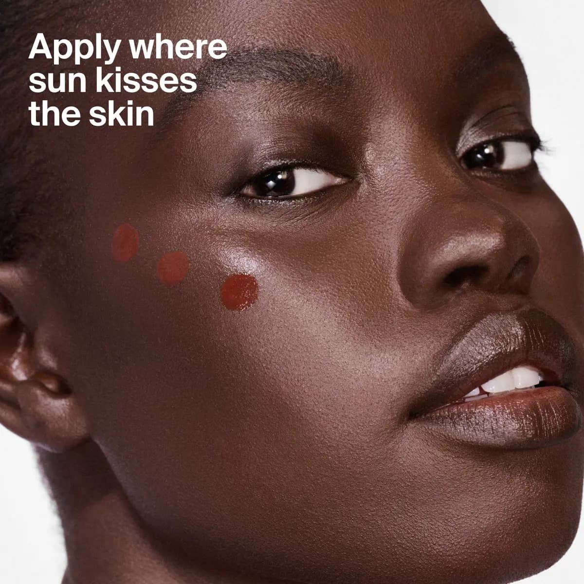 Deep-skinned model with three reddish-brown bronzer dots on the cheekbone and text 'Apply where sun kisses the skin