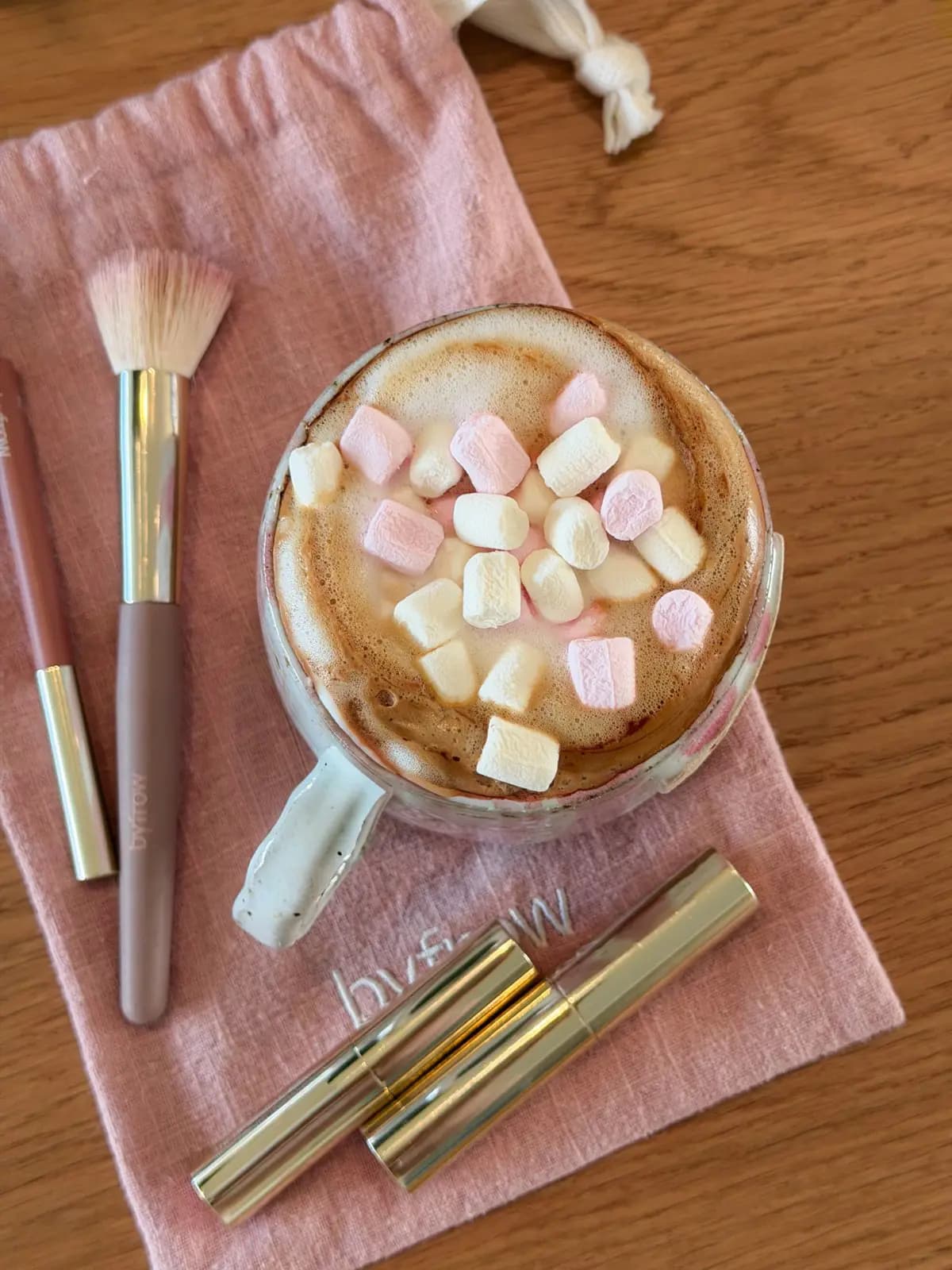Byfrow brush, pencil and gold tubes on a pink embroidered drawstring bag beside hot chocolate with marshmallows