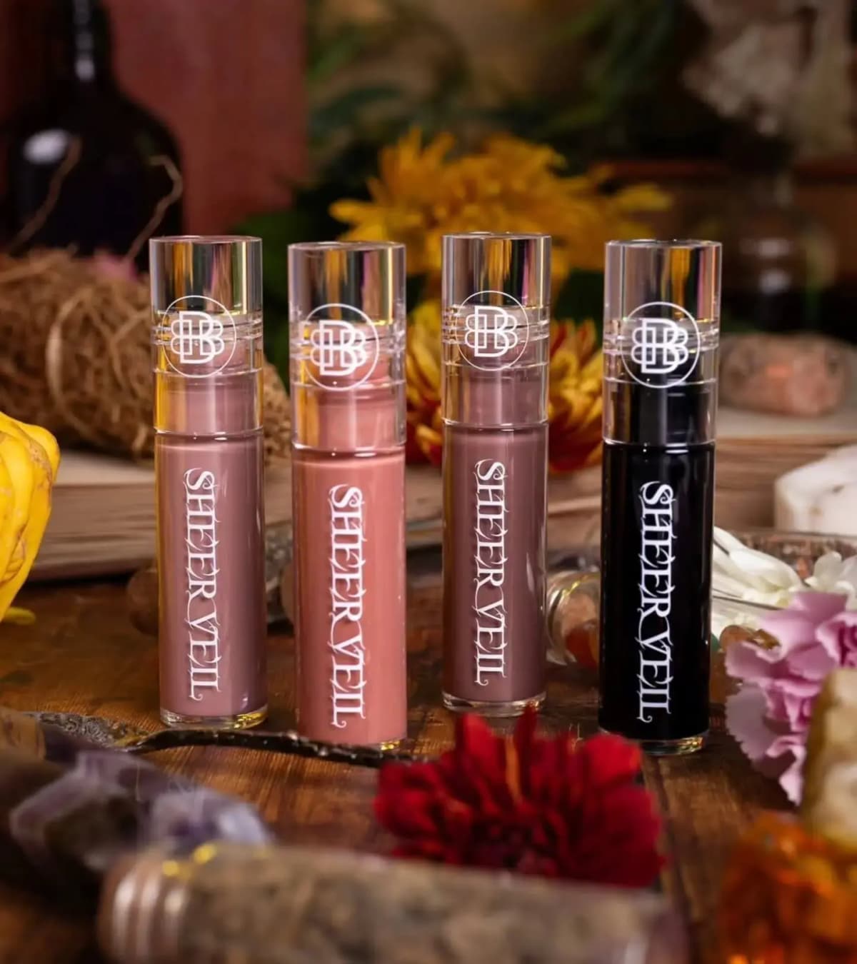 Four Bella Beauté Bar Sheer Veil Lipgloss tubes, three in mauve and rosy nudes and one black, among flowers on a wooden table