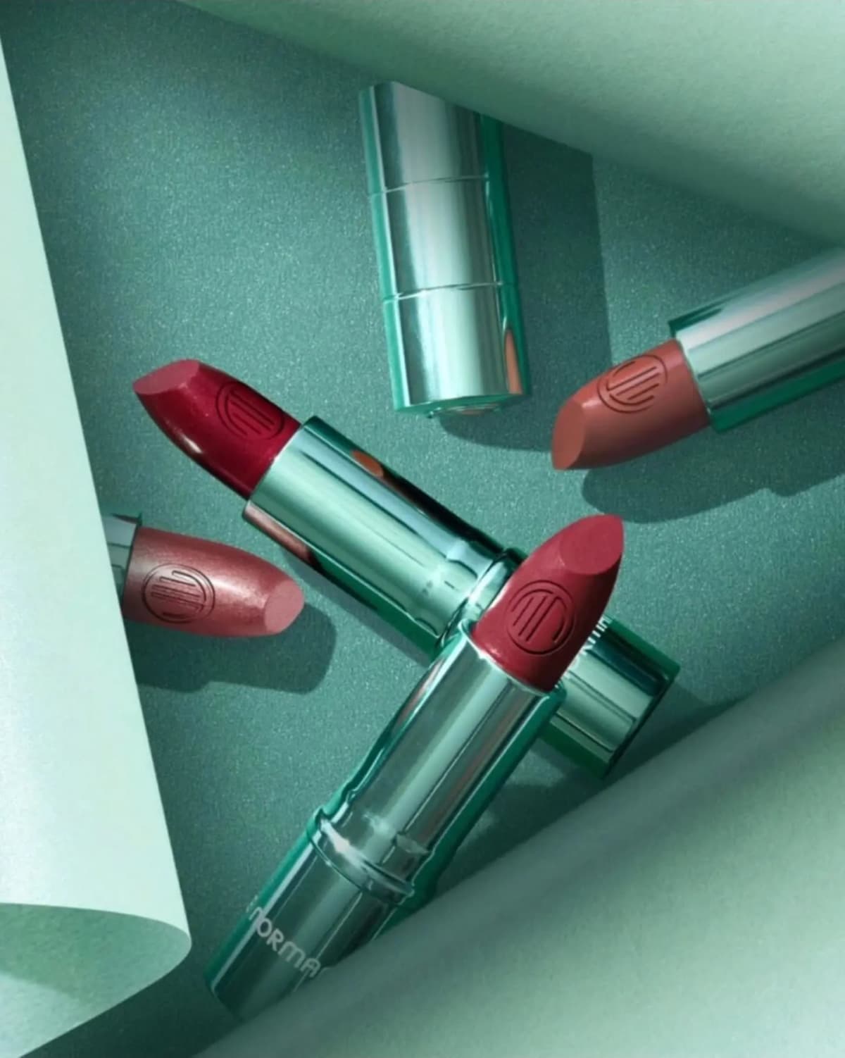 Four red, berry, pink and nude lipsticks in shiny green cases with embossed logos on a teal textured backdrop