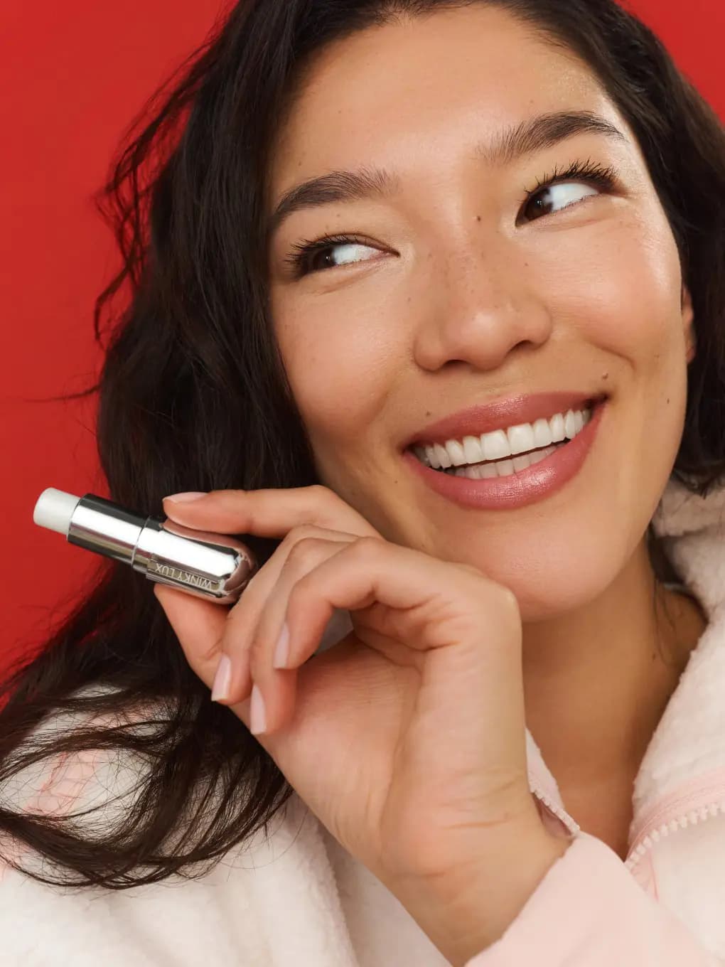 Smiling model with glossy rosy lips holding the silver Winky Lux lip balm against a red background
