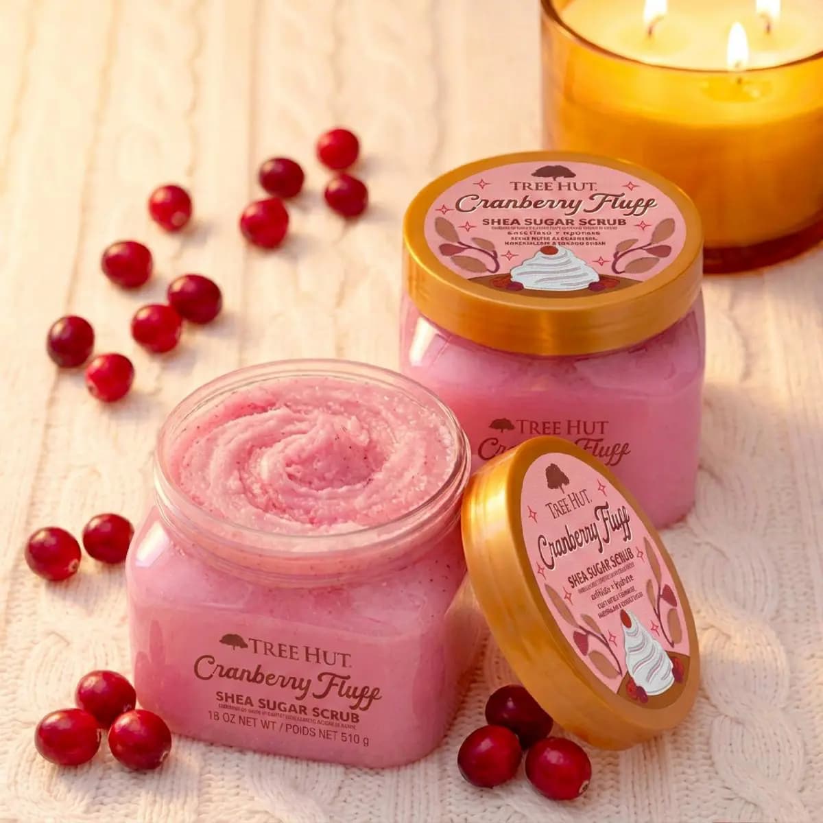 Two pink Cranberry Fluff Shea Sugar Scrub jars beside fresh cranberries and a lit amber candle on knit fabric