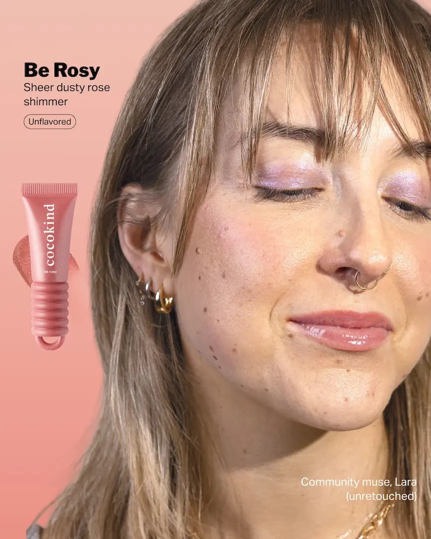Model with bangs and nose rings wearing Be Rosy, a sheer dusty rose shimmer, beside its pink tube