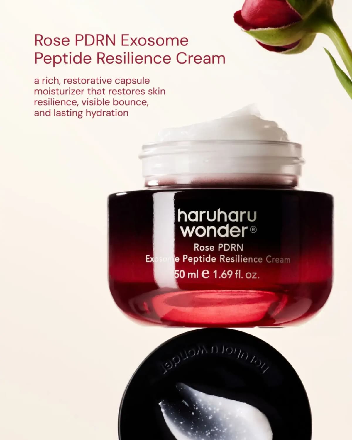 Open red gradient jar of white cream above a black lid holding a glossy swatch, beside a red rosebud on cream backdrop