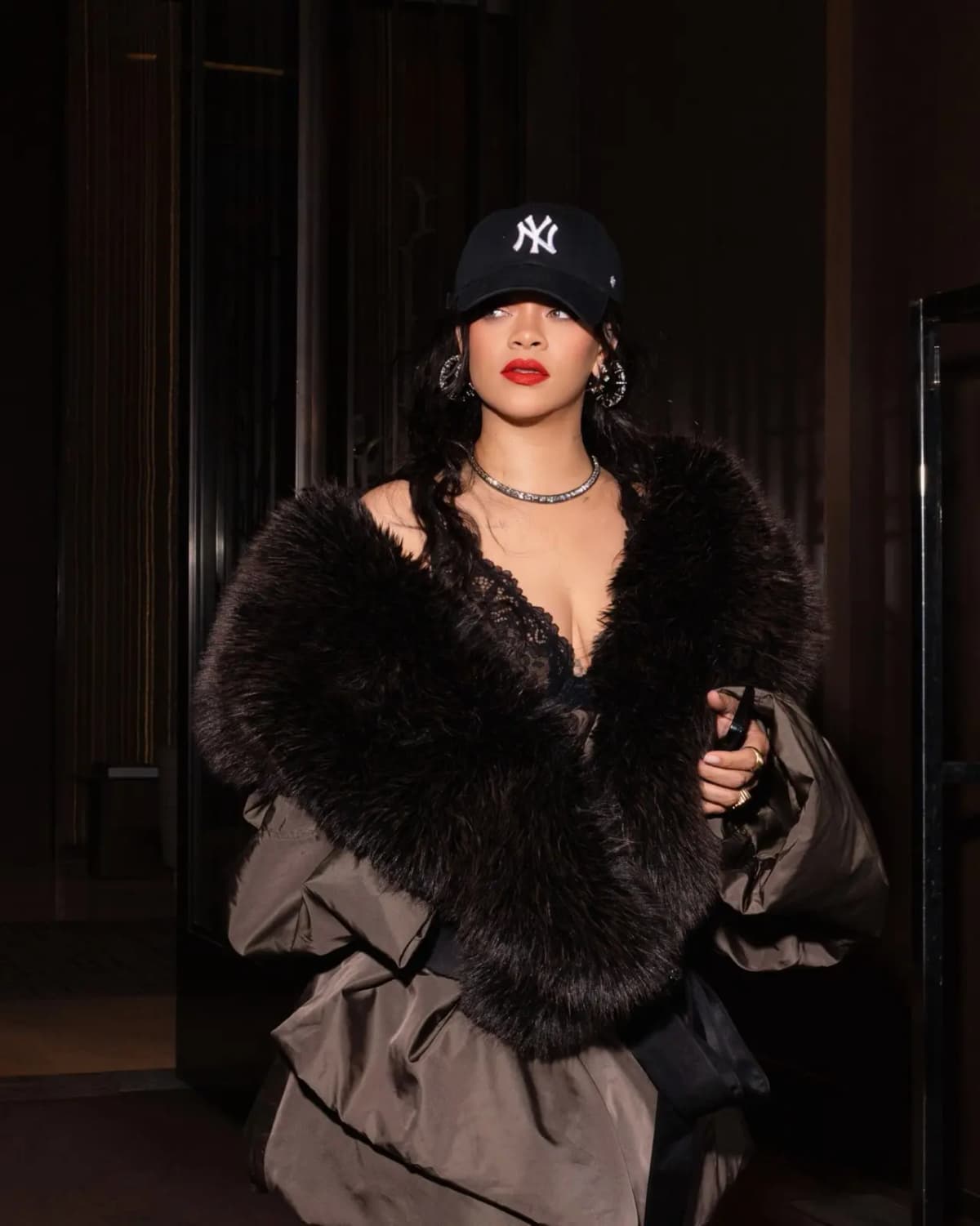 Rihanna in a black NY cap, faux fur stole and lace top wearing bright red matte lips while stepping out at night