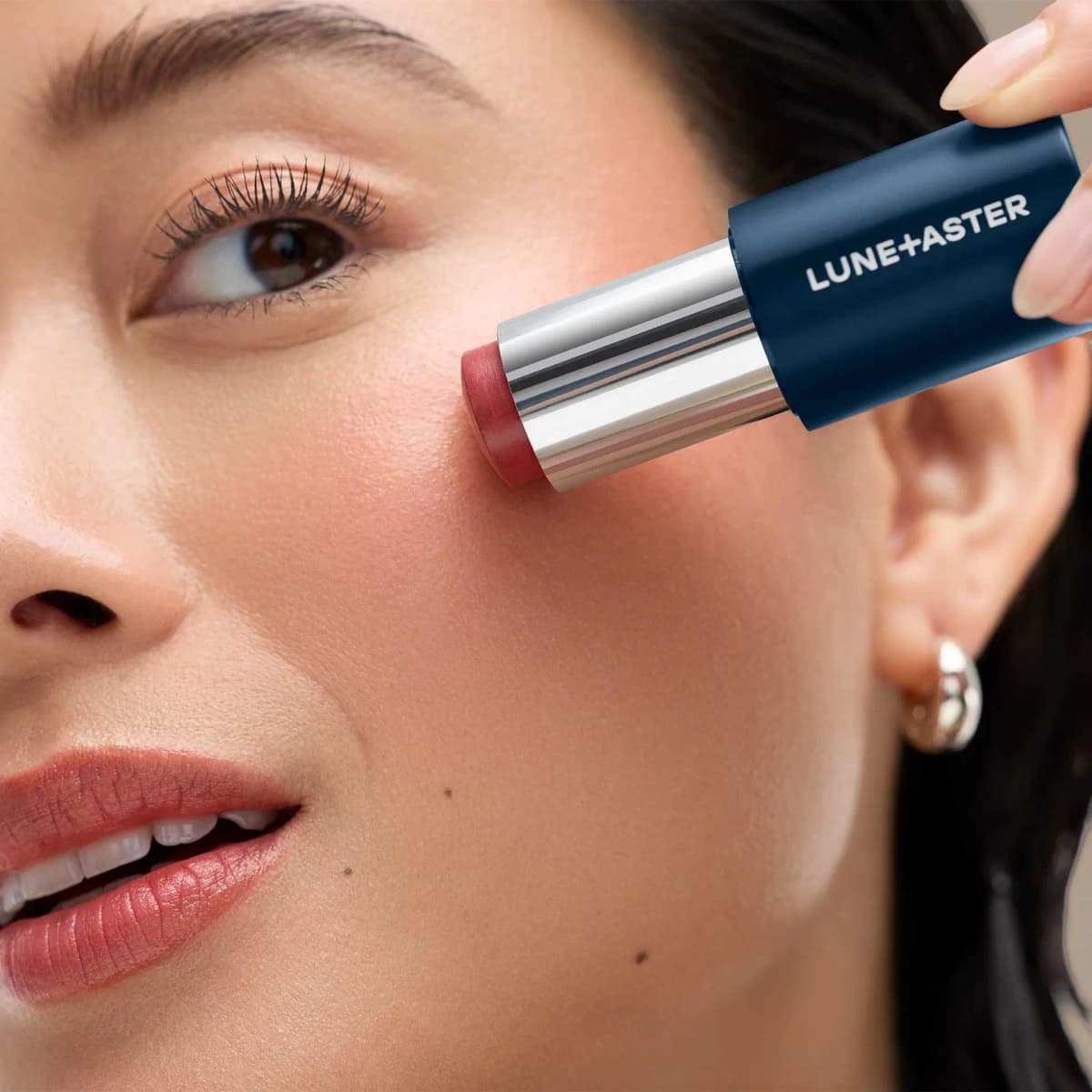 Model applying a berry-rose cream stick to the cheek, with a matching rosy lip
