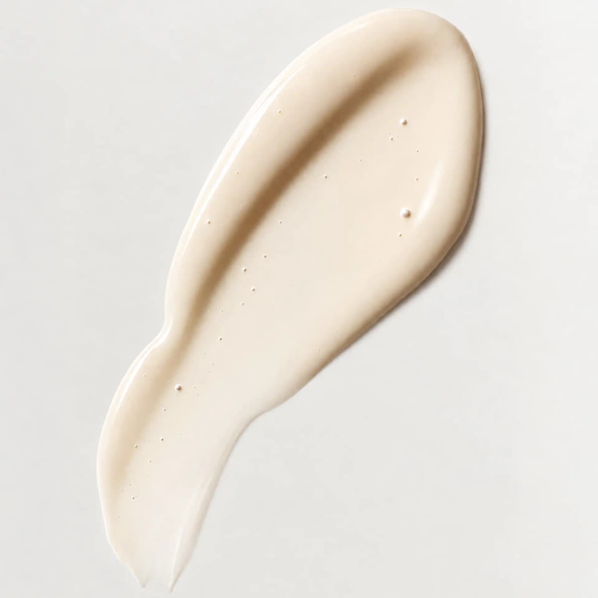 Close-up swatch of glossy ivory serum smeared across a pale background, dotted with tiny bubbles