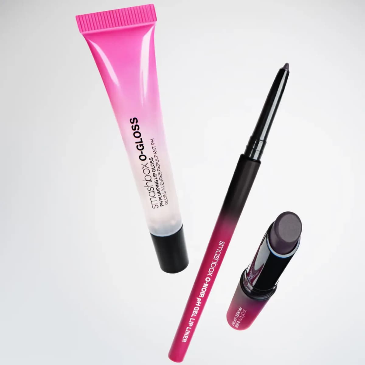 O-Gloss tube floating beside a Smashbox O-Noir pH gel lip liner and a dark plum stick on a pale grey backdrop