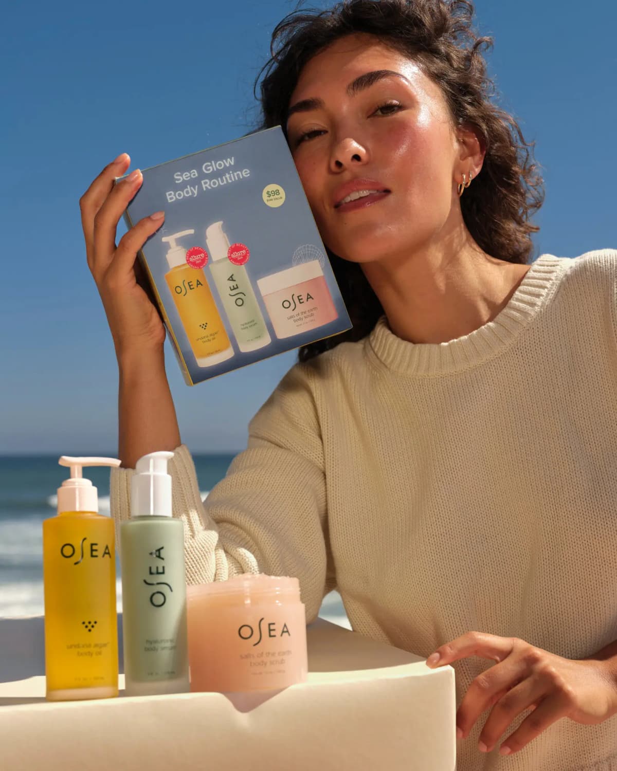 Model in a cream sweater holds the $98 Sea Glow Body Routine box by the beach, with the three products in front