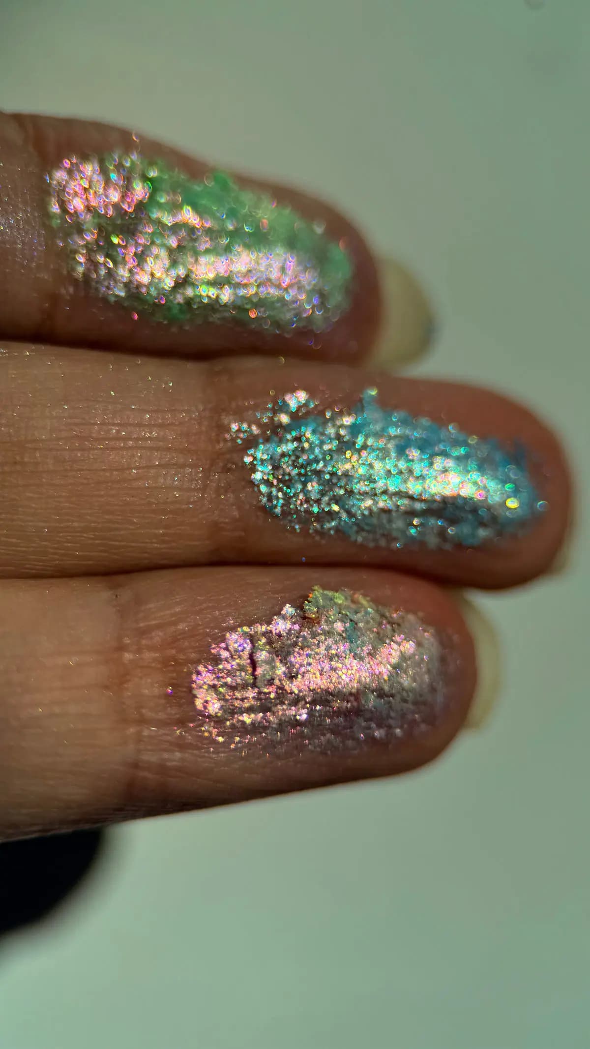 Close-up finger swatches of green, aqua and pink-gold iridescent glittery eyeshadows