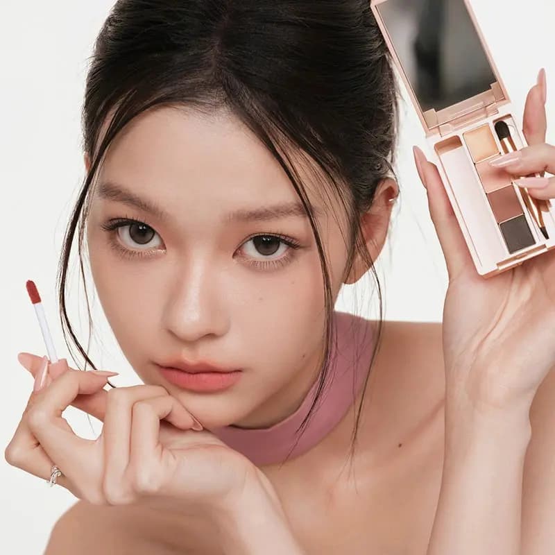 Model with soft natural makeup holds an open pink JUDYDOLL palette in one hand and a red lip tint wand in the other.