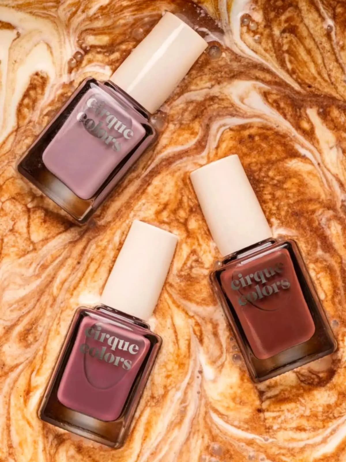 Three polish bottles in lilac, mauve and brown lying in swirled milky coffee