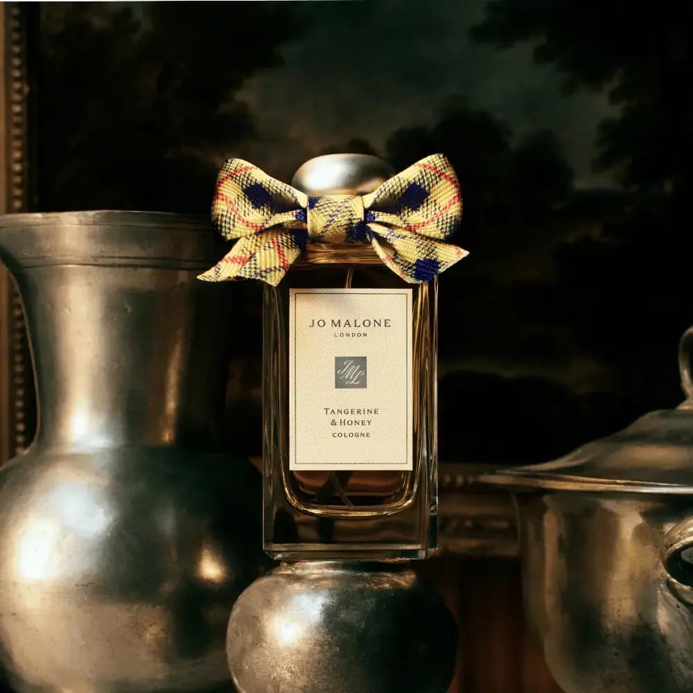 Cologne bottle with tartan bow perched on a pewter jug among silver vessels, set against a dark landscape painting