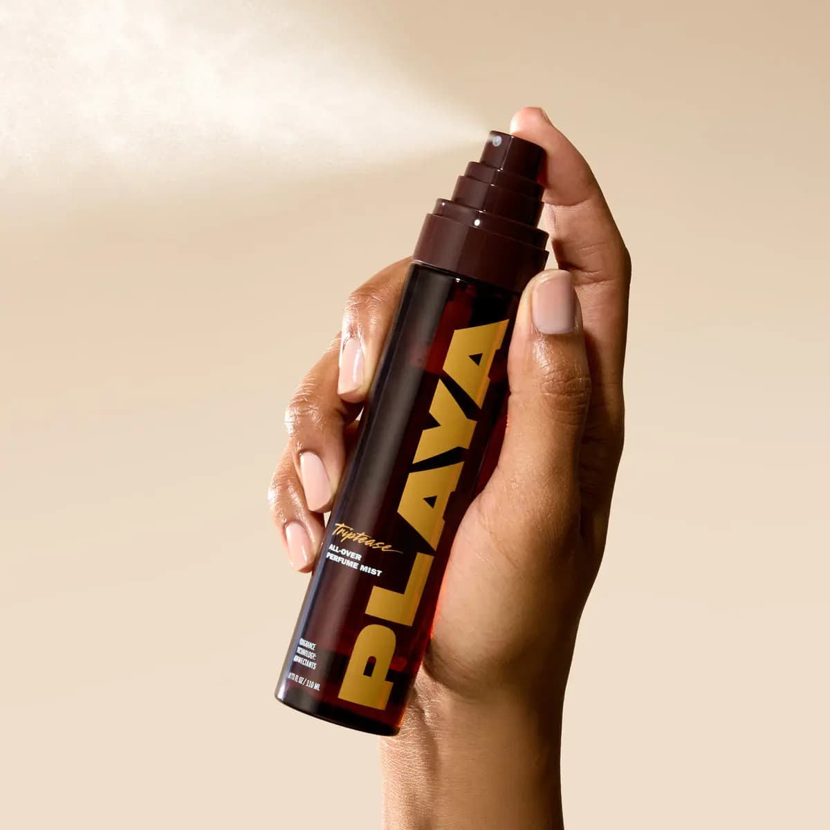 Hand spraying the brown and gold mist bottle, a fine cloud of fragrance drifting against a beige backdrop
