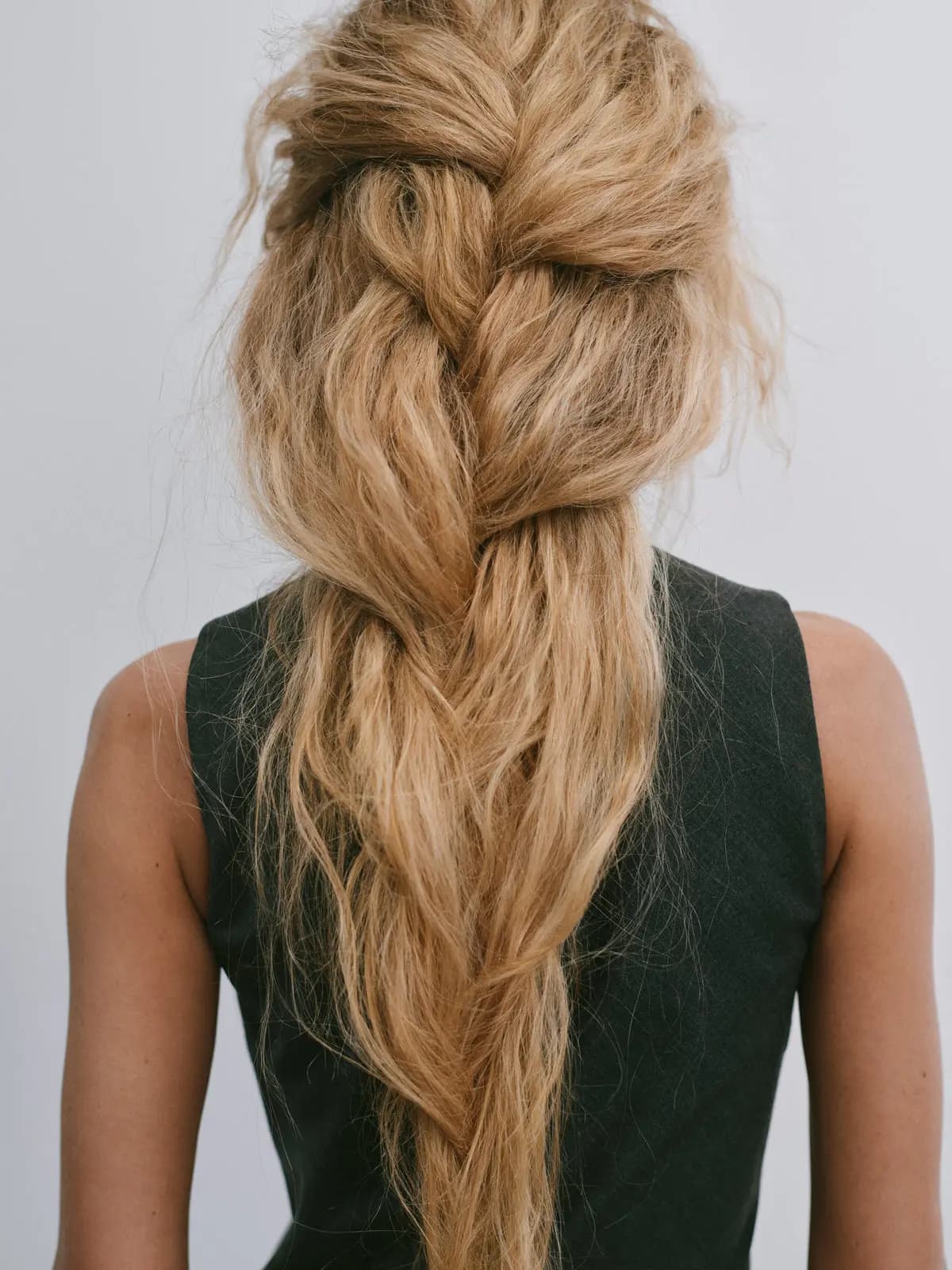 Back view of a loose, textured honey-blonde braid falling over a dark grey sleeveless top