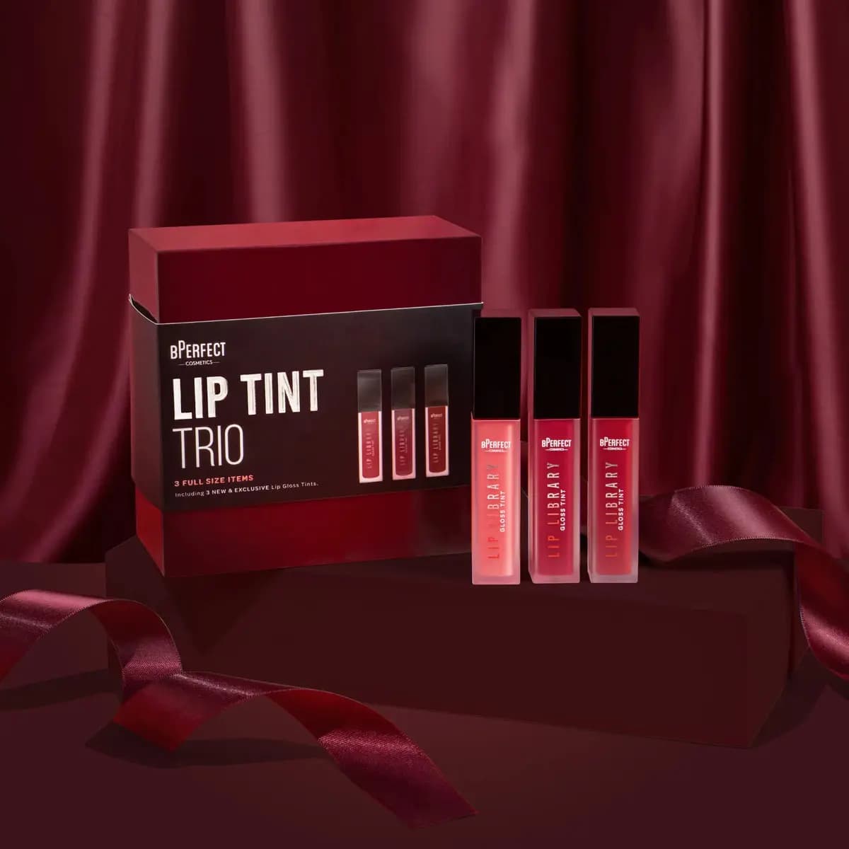 BPerfect Lip Tint Trio box beside three Lip Library gloss tints in coral, rose and red on a burgundy stand