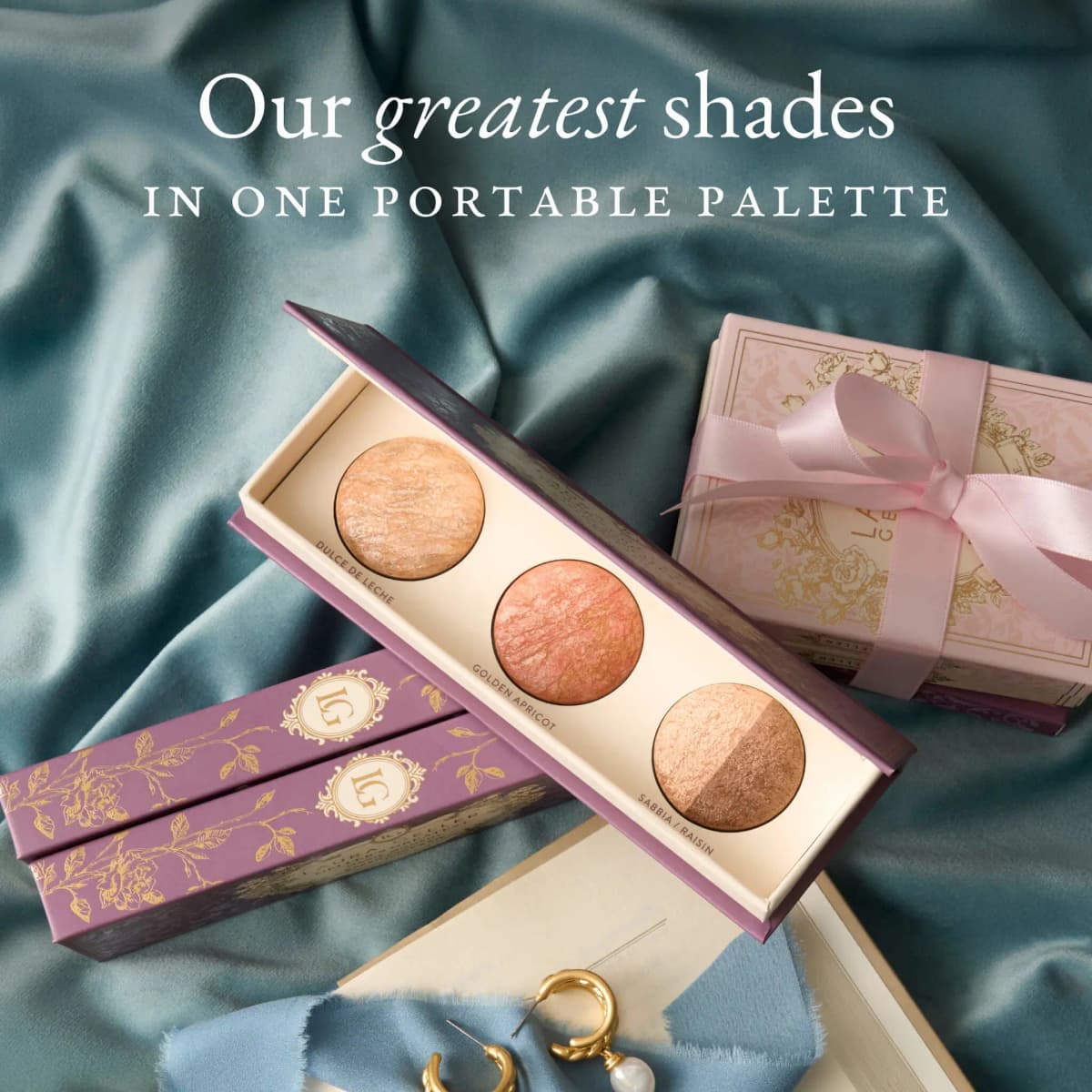Open Magic Moment palette on teal satin with purple cases, a pink ribboned gift box and gold earrings; portable palette text.