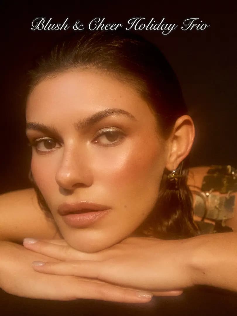 Blush & Cheer Holiday Trio campaign: dark-haired model with gold eyeshadow, glowing cheeks, chin resting on hands