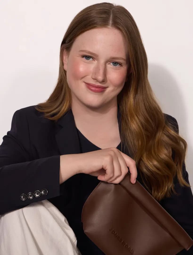 Smiling auburn-haired model in navy blazer with rosy cheeks and lips holds the brown Jones Road clutch