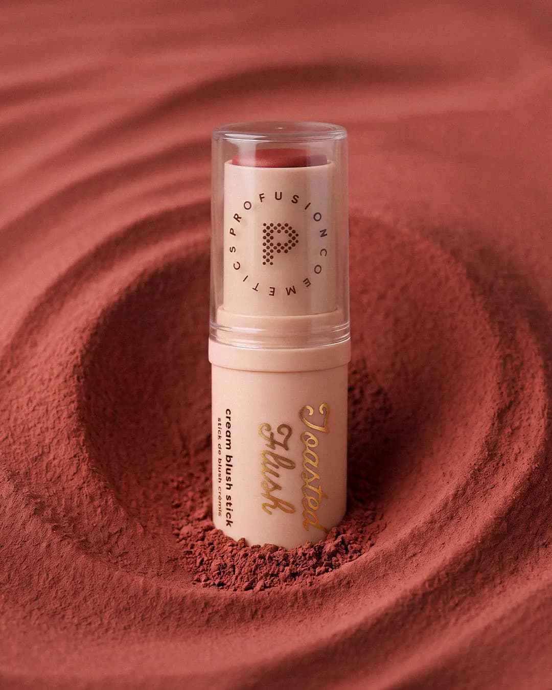 Toasted Flush cream blush stick standing in a swirl of rosy terracotta powder.
