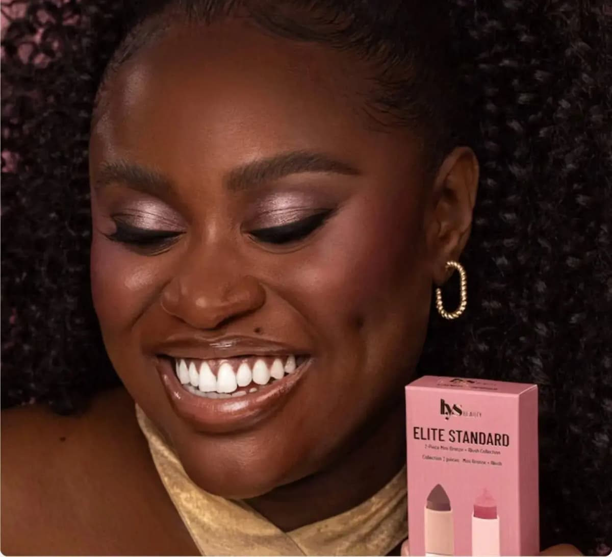 Smiling model with shimmery mauve eyeshadow and curly hair holds a LYS Beauty Elite Standard box