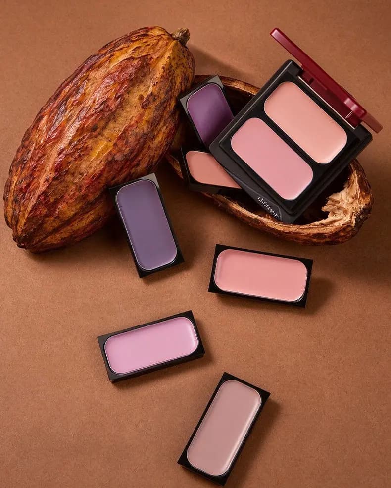 Duo compact and five cream blush pans in mauve, peach, lilac and nude arranged around cocoa pods on a tan backdrop