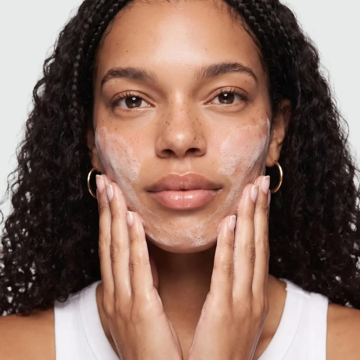 Curly-haired model with freckles massaging white Shed the Day Cleanser foam over the cheeks and chin