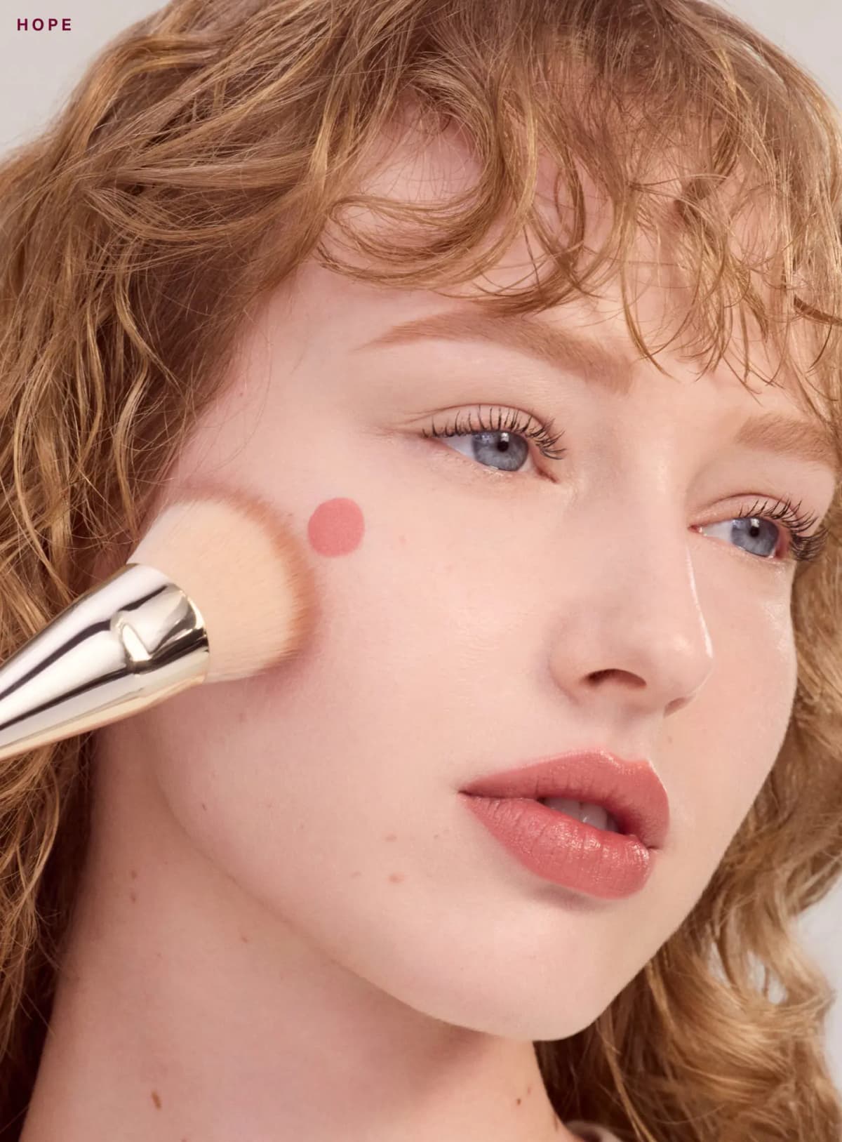 Red-haired model with a dot of Hope blush on her cheek being blended with a gold-ferruled brush