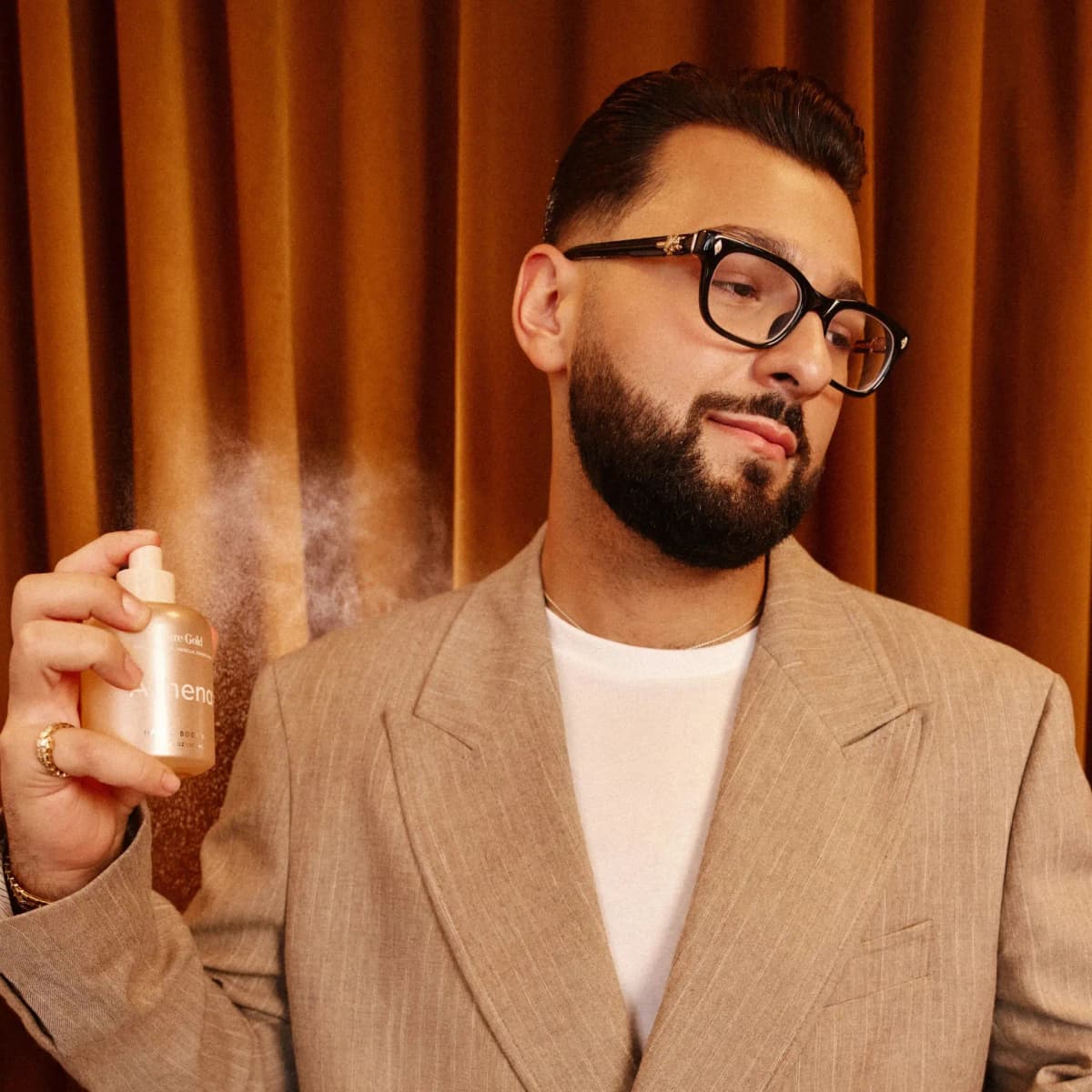 Paul Reacts in glasses and a pinstriped beige blazer spritzing the Bare Gold mist in front of an amber velvet curtain