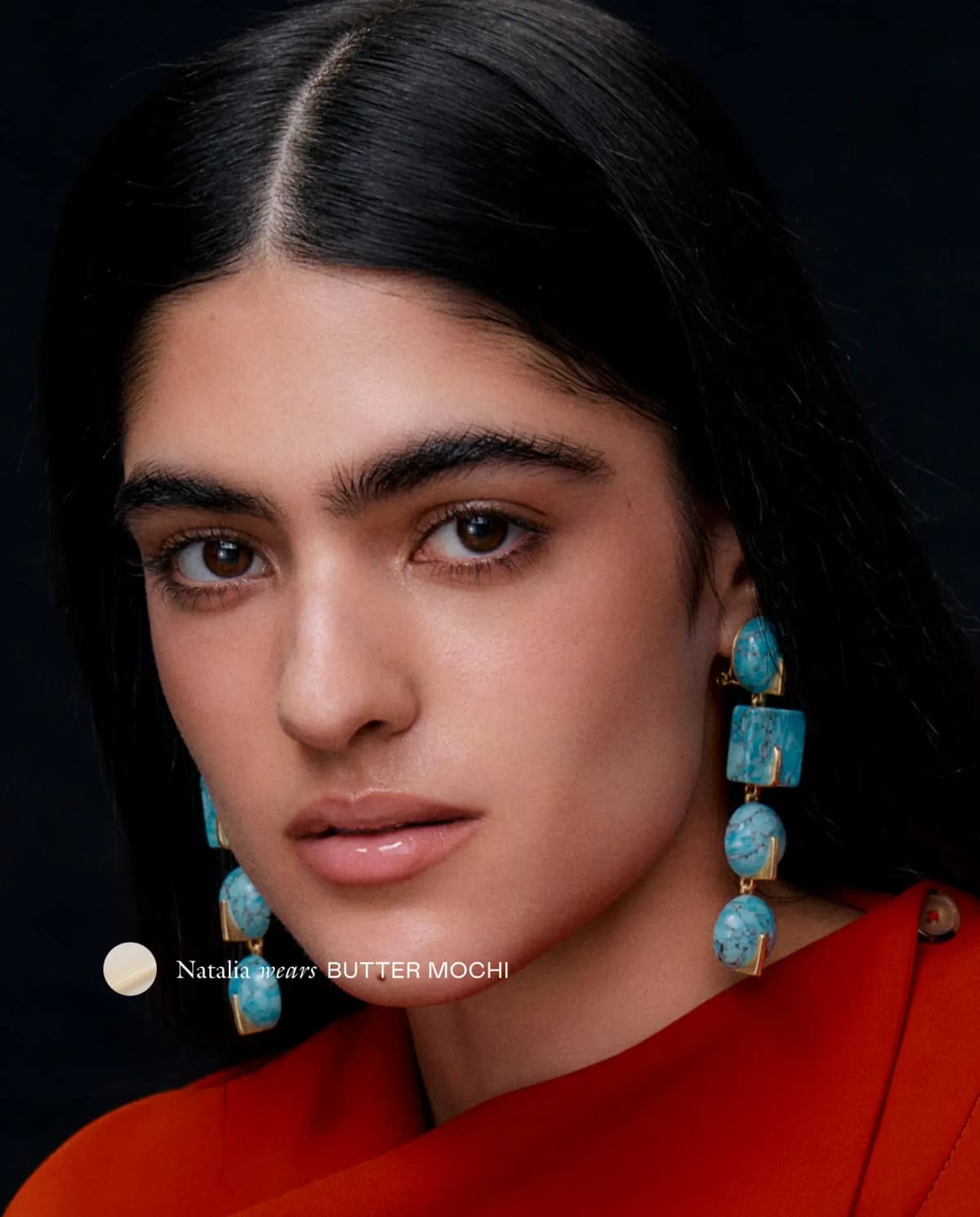 Natalia in turquoise drop earrings and a red top wears sheer, glossy Butter Mochi