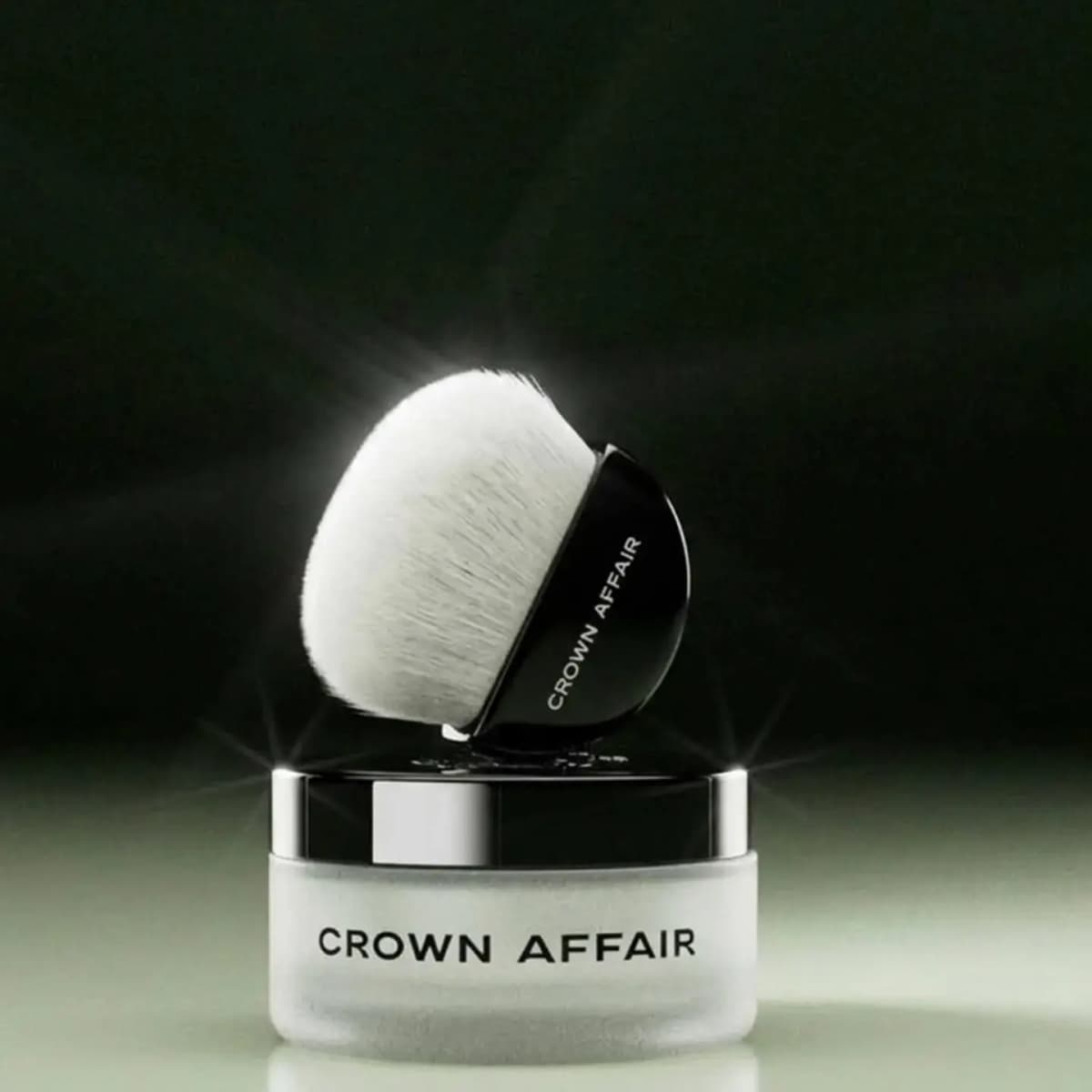 Crown Affair frosted jar with silver lid topped by a domed white brush with black handle, glowing on a dark green backdrop