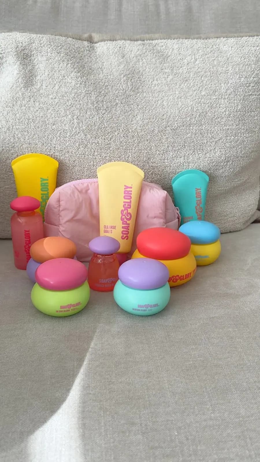 Soap & Glory skincare collection of tubes, jars and bottles in bright colours with a pink pouch on a beige sofa