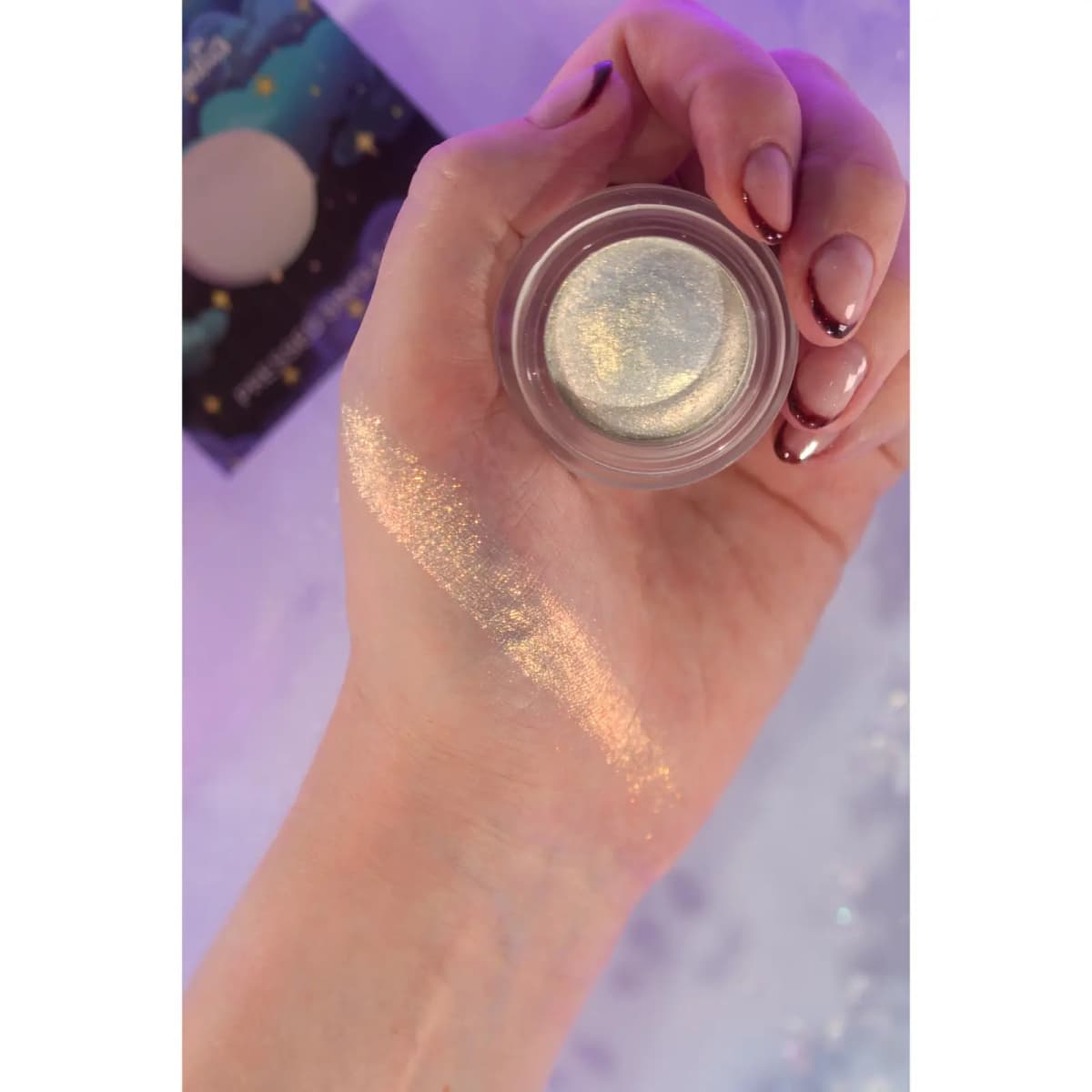 Hand holding a jar of pearly gold-green shimmer cream with a sheer golden-pink swatch across the back of the hand