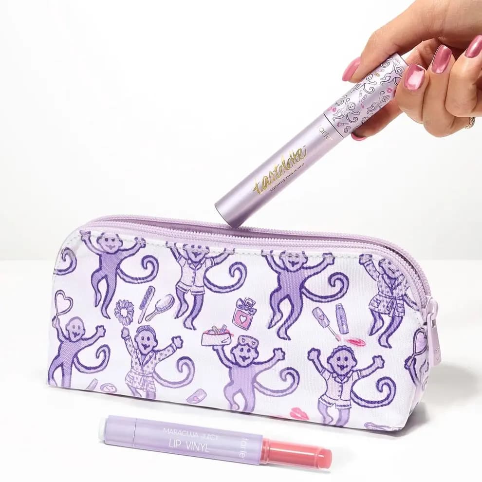Hand placing tartelette mascara into a lavender monkey-print zip pouch, Lip Vinyl lying in front