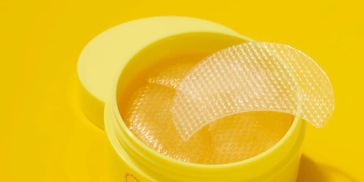 Open yellow jar of golden textured gel patches with one clear patch draped over the rim on a bright yellow backdrop