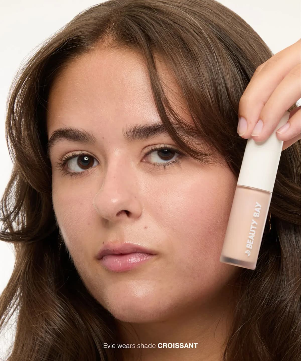 Model with long brown hair holds a light Beauty Bay concealer bottle to her cheek; caption says she wears Croissant