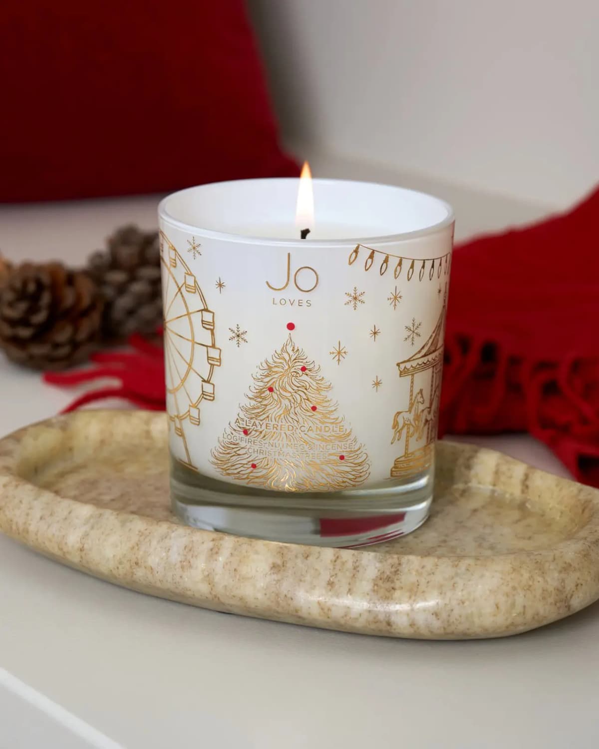 Lit Jo Loves Layered Candle in white glass with gold Christmas fair print on a stone tray, pine cones and red throw behind