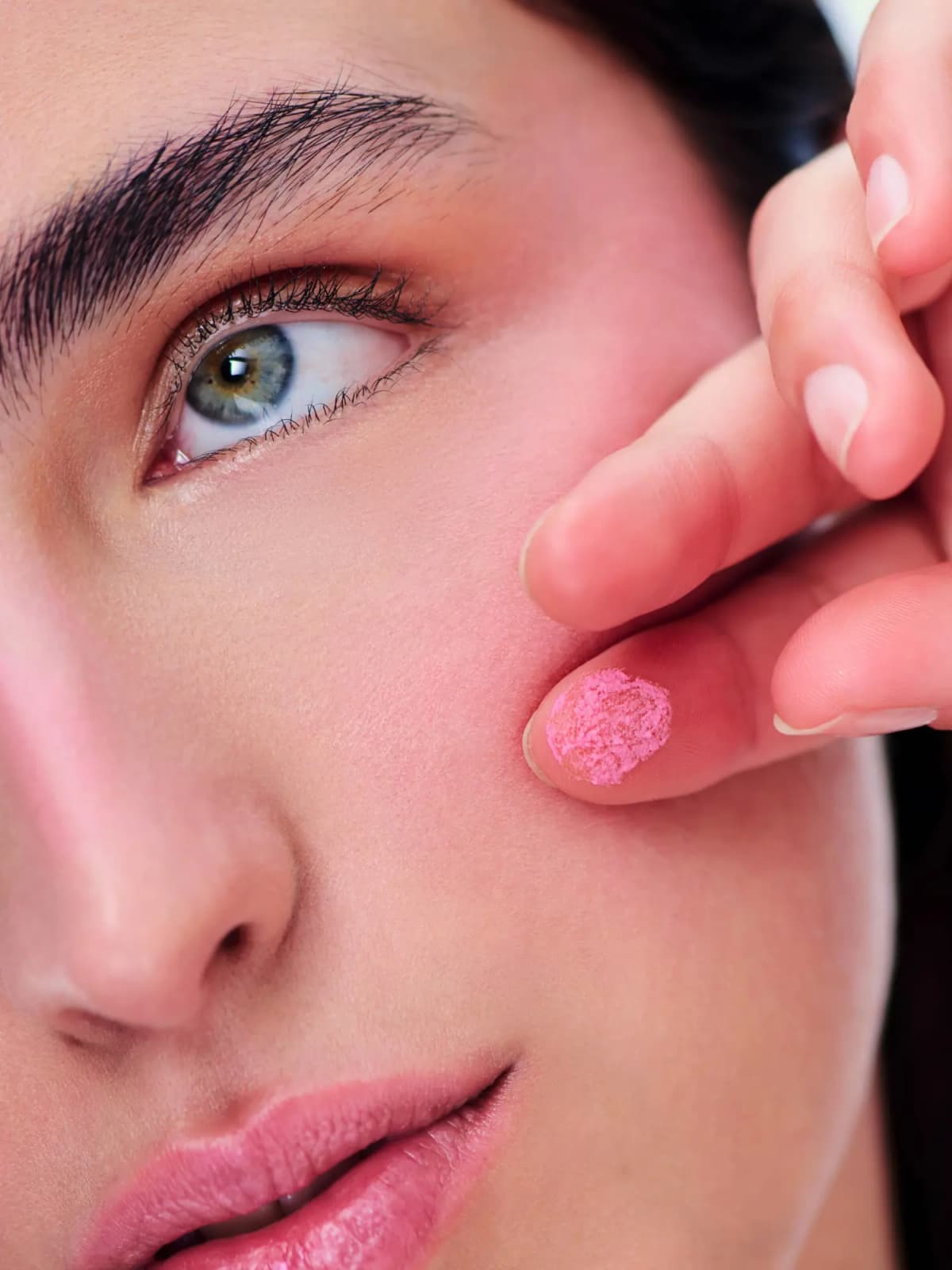 Close-up of a model's flushed cheek with a fingertip dabbed in powdery pink mousse blush